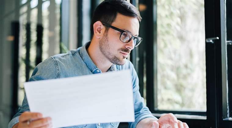 Serious pensive thoughtful focused young casual business accountant bookkeeper in office looking at and working with laptop and income tax return papers and documents