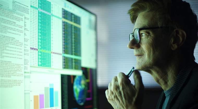 A mature man working in his home office. He’s studying a large computer monitor displaying a variety of numerical data, global information