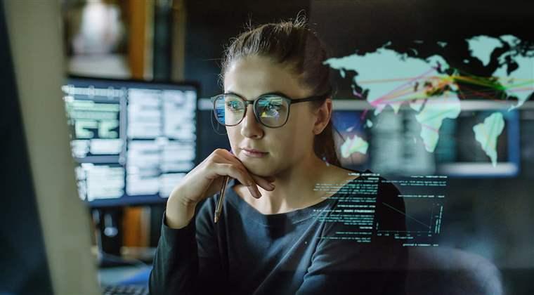 A young woman, wearing glasses, surrounded by computer monitors in a dark office. In front of her there is a see-through displaying showing a map of the world with some data.