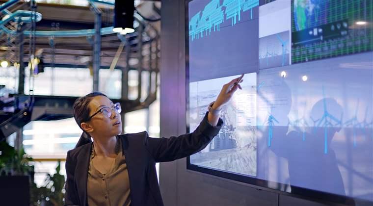 A young Asian woman conducting a seminar / lecture with the aid of a large screen. The screen is displaying data & designs concerning low carbon electricity production with solar panels & wind turbines. These are juxtaposed with an image of conventional fossil fuel oil production.