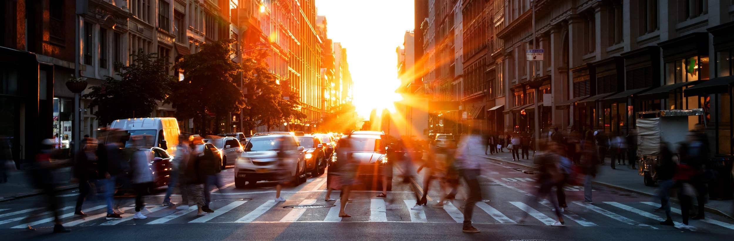 Crowds of busy people walking through the intersection of 5th Avenue and 23rd Street in Manhattan, New York City with bright sunset background