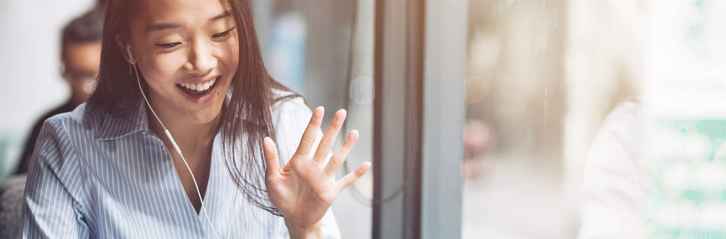 Portrait of a young asian freelancer in earphones looking at the screen of her laptop and waving while making a video call in the cafe. Horizontal shot. Front view