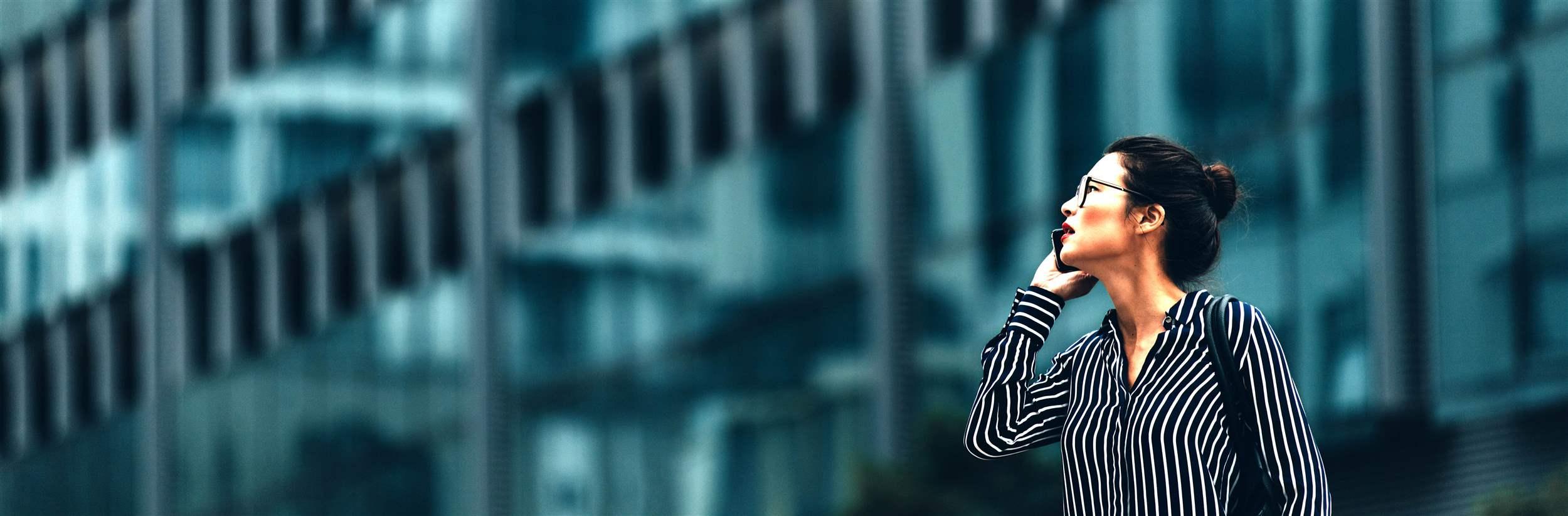 Businesswoman walking outdoors and talking on mobile phone. Female business professional walking outside with an office building in background.