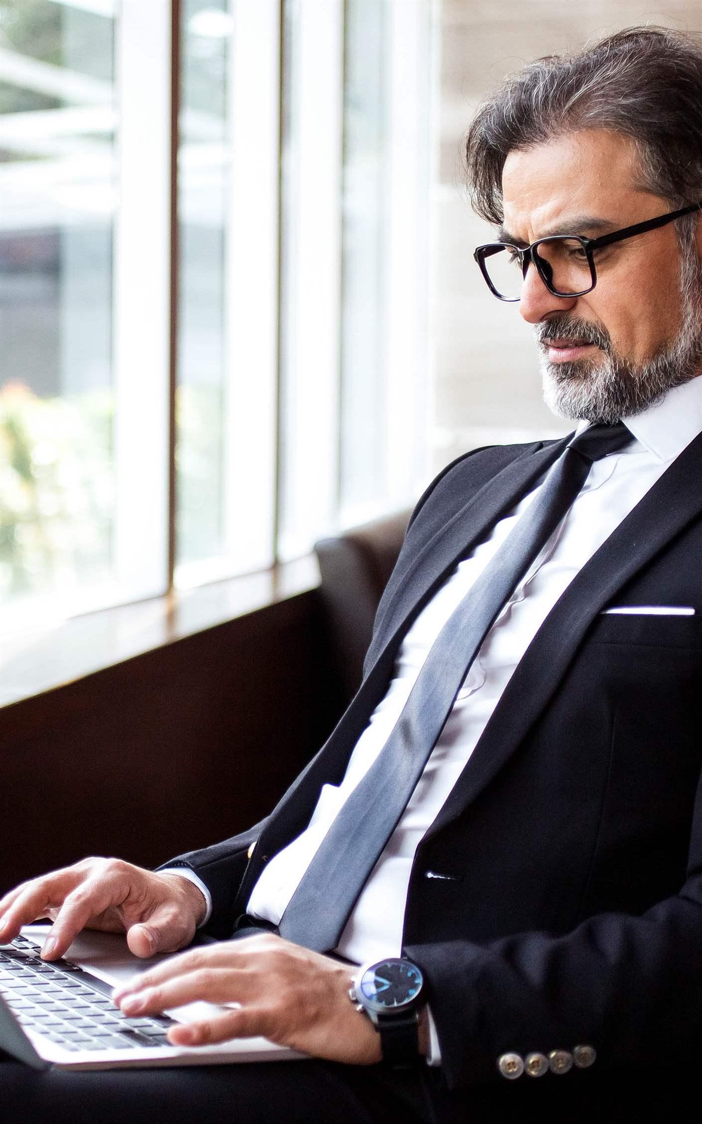 Concentrated senior businessman using laptop in lobby and examining report. Serious manager immersed in work out of office. Technology in business concept