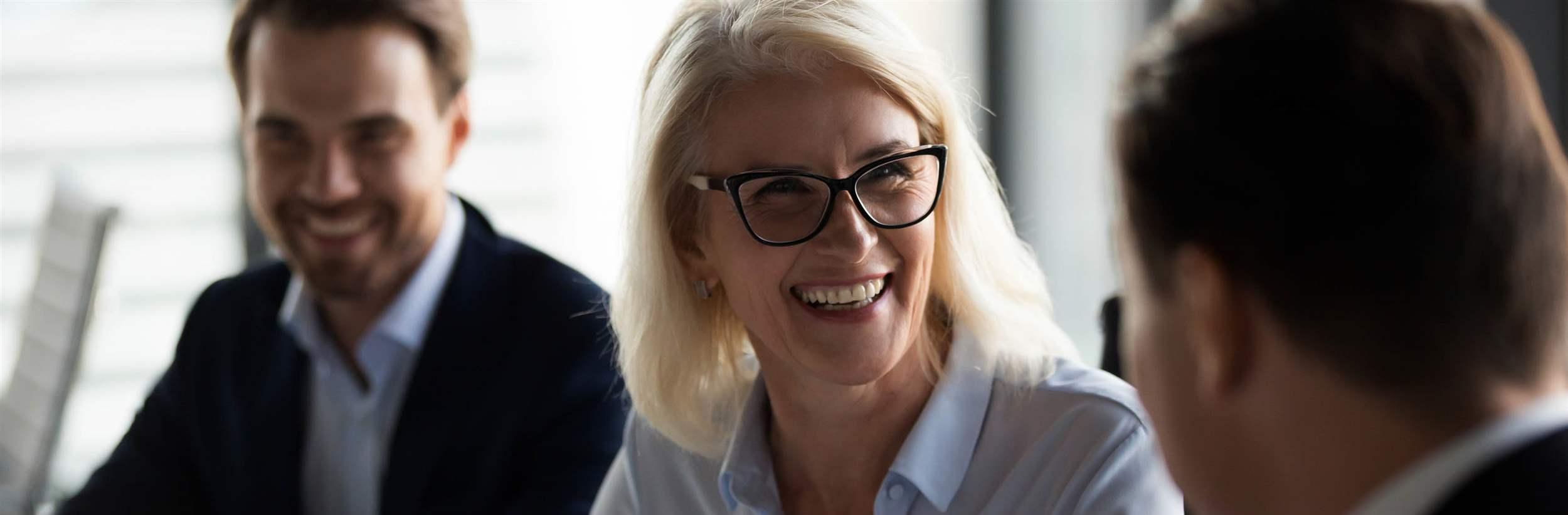 Friendly middle aged female leader laughing at group business meeting, happy old businesswoman enjoying fun conversation with partner, smiling mature business coach executive talking to colleague