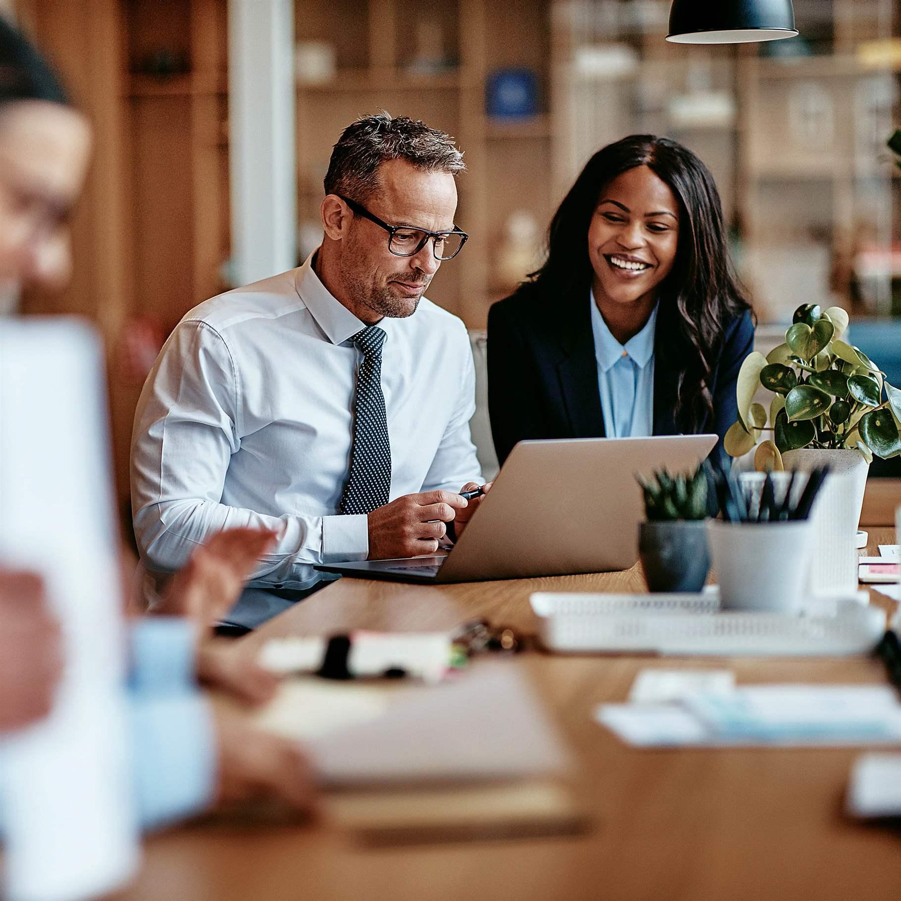 Two diverse businesspeople smiling while working on a laptop together at the end of a boardroom table in an office