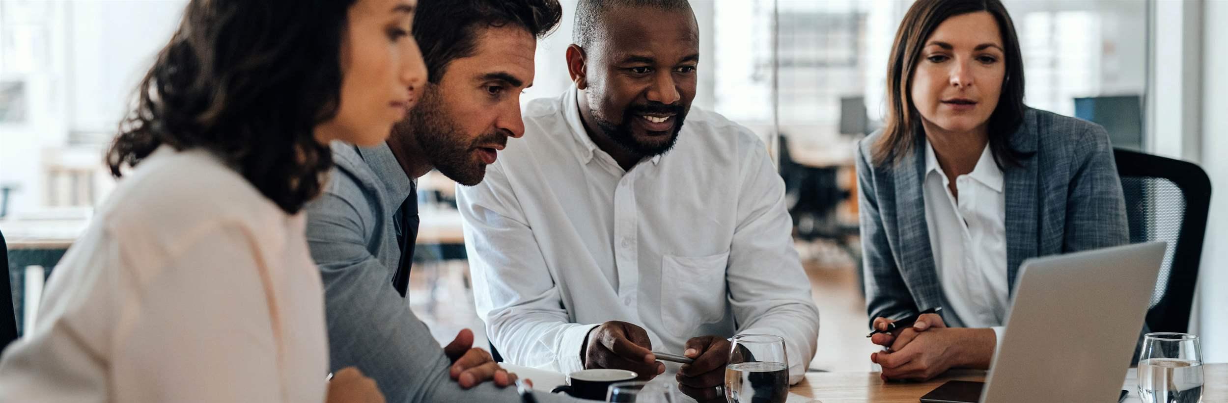 Diverse group of smiling businesspeople working together over a laptop during a meeting in the boardroom of a modern office