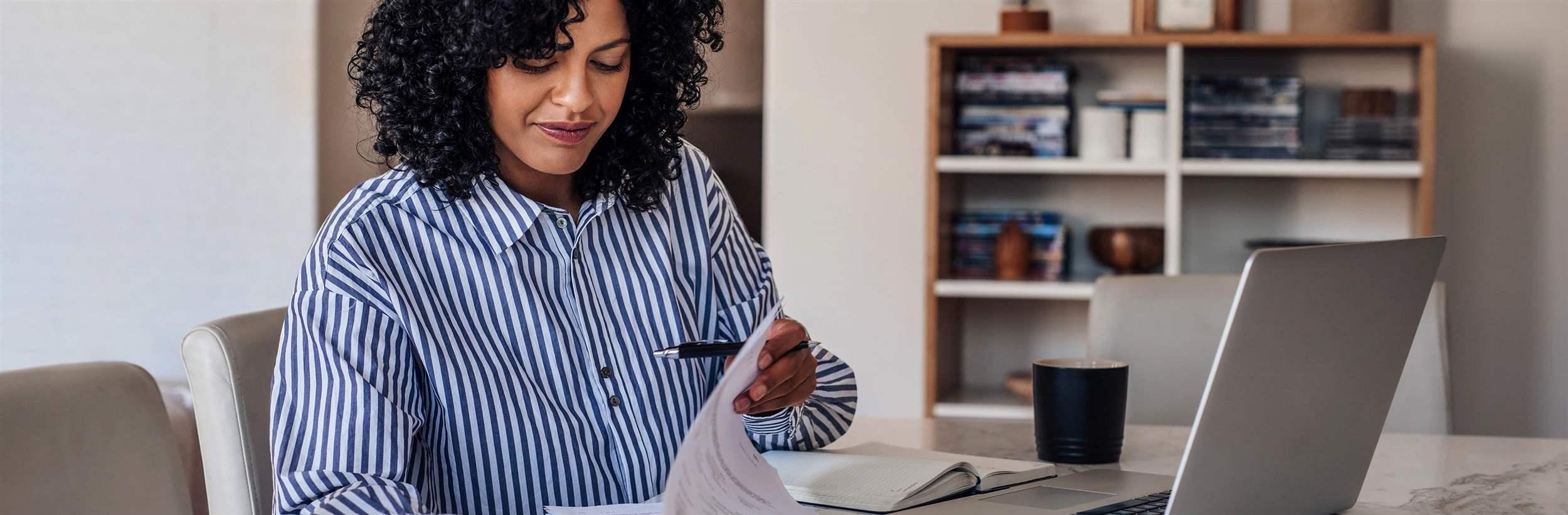 Smiling young female entrepreneur going through paperwork while working on a laptop at her dining room table at home