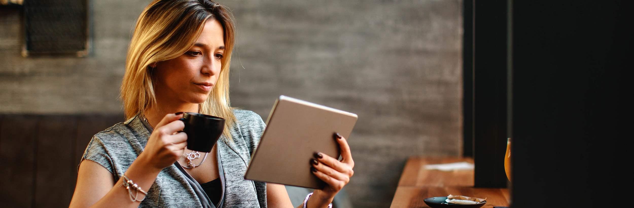Young beautiful woman relaxing in coffee shop reading electronic book via internet on digital tablet and drink coffee