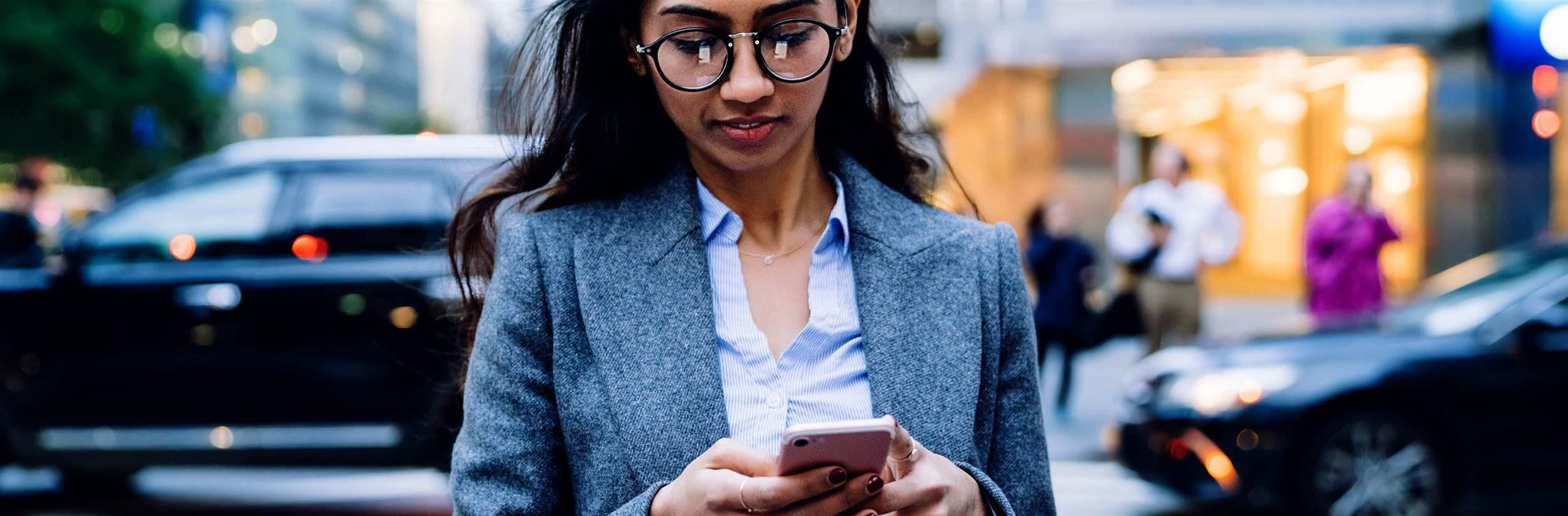 Asian business woman in glasses and gray jacket browsing in mobile phone while crossing road in New York City