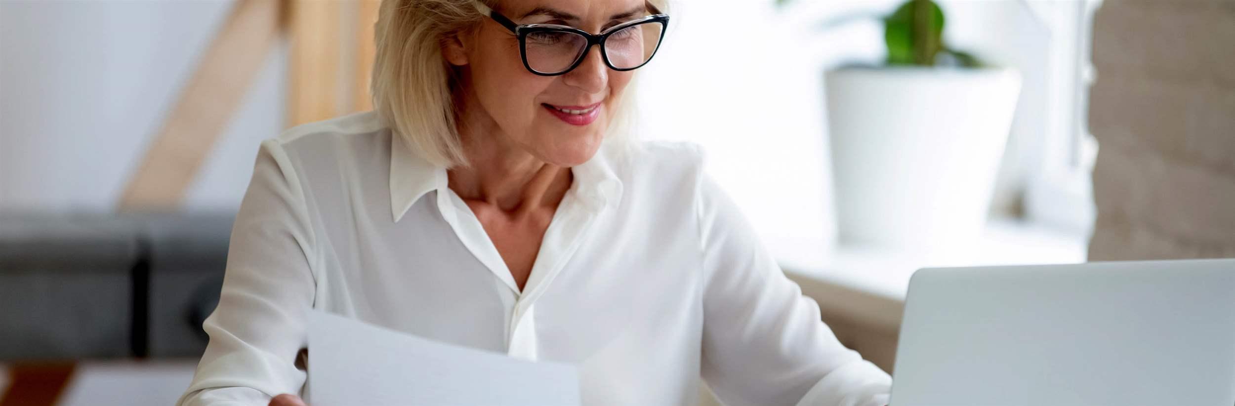 Aged businesswoman typing on pc holds paper prepare report analyzing work results feel satisfied do paperwork use business app online software for data analysis, financier manage firm finances concept