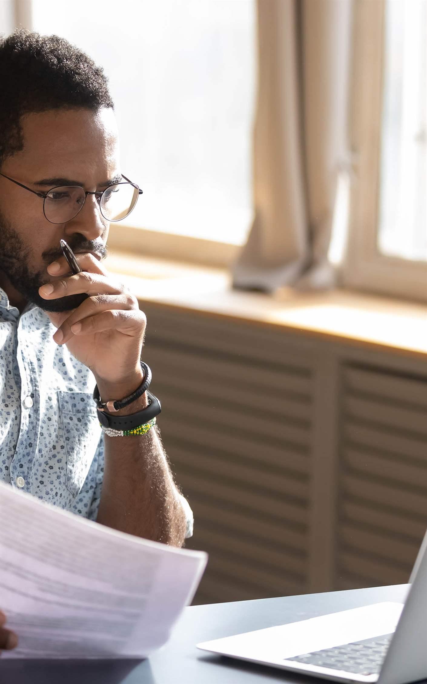 Focused serious businessman accountant analyst holding documents looking at laptop computer screen doing online trade market tech research thinking working sit at home office desk