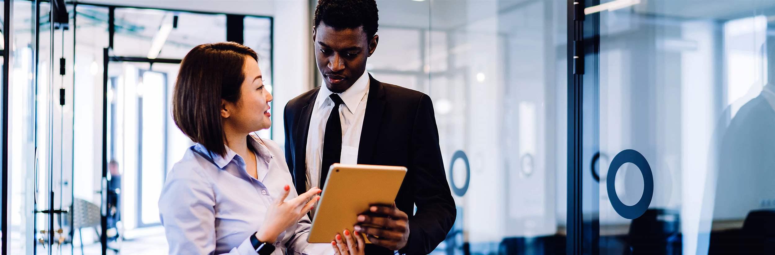 Multiracial young couple in formal wear browsing tablet while female explaining interesting information standing in corridor of company office