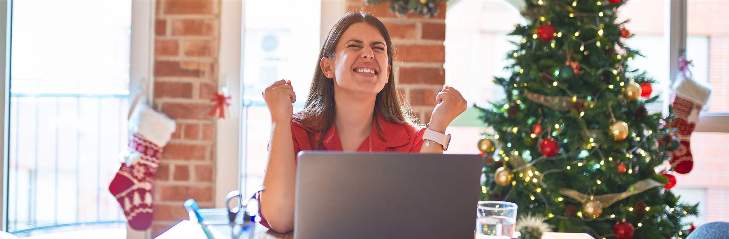 Beautiful woman sitting at the table working with laptop at home around christmas tree celebrating surprised and amazed for success with arms raised and eyes closed. Winner concept.