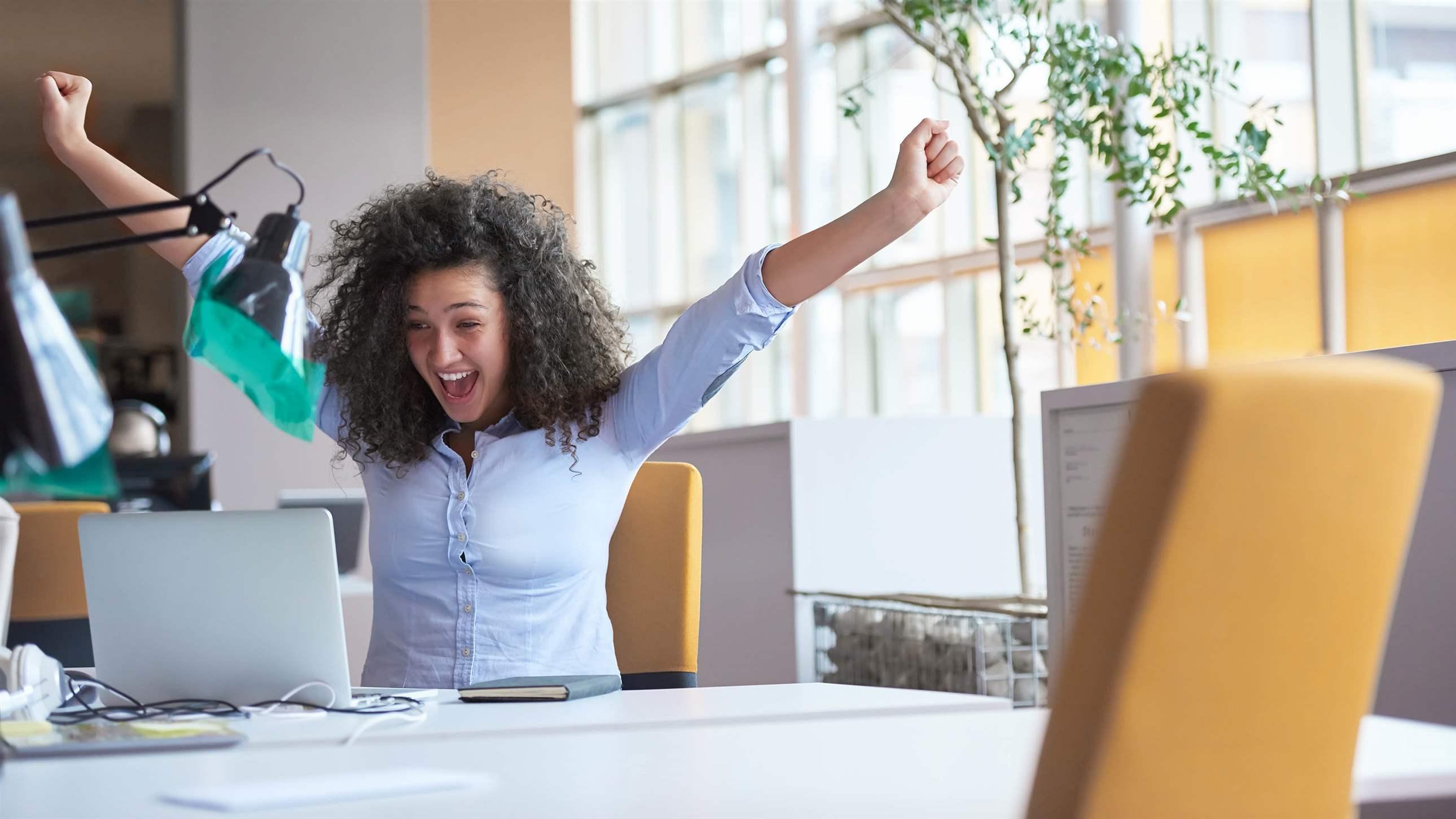 Woman in office at laptop happy with arms raised in excitement