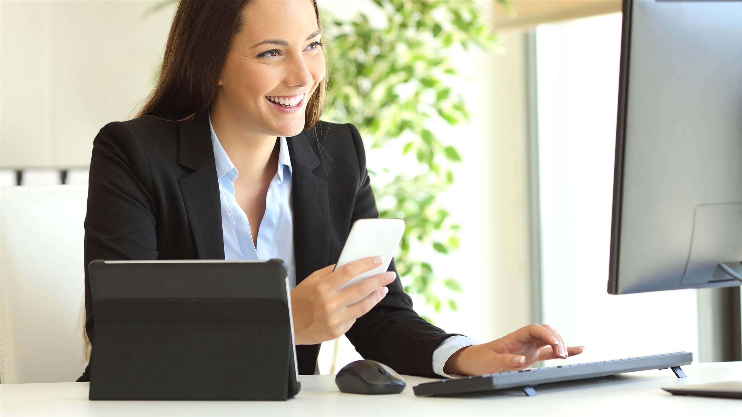 Happy businesswoman working on line with multiple devices and watching a desktop computer at office