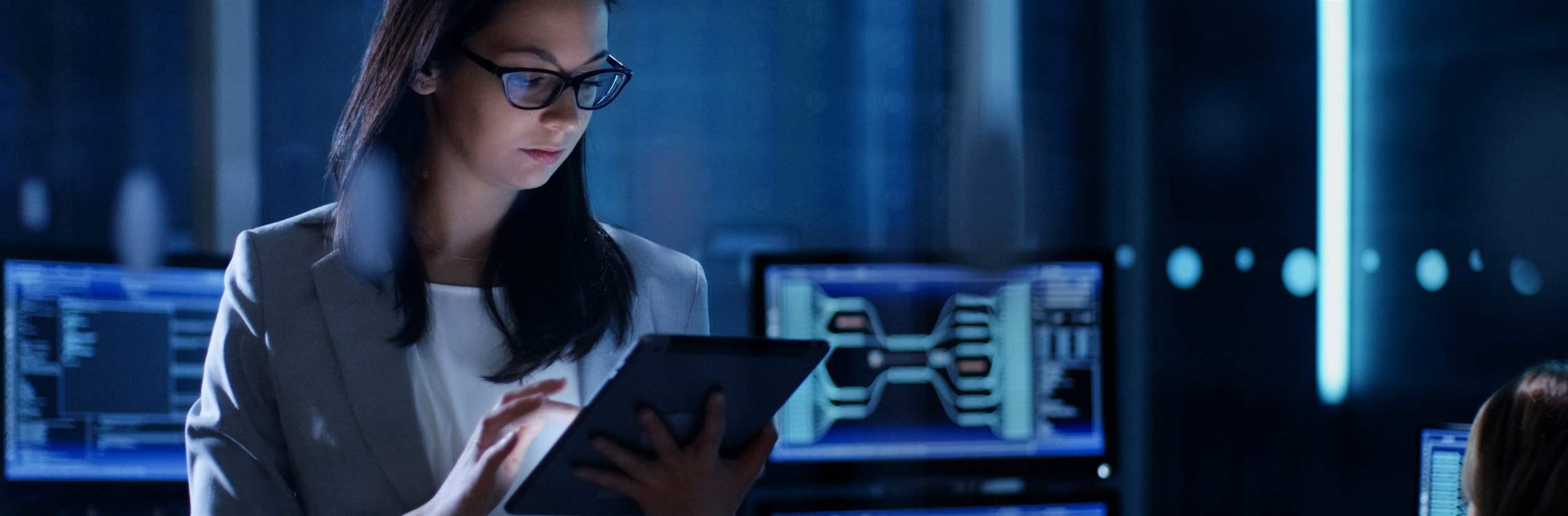 Young Female Government Employee Wearing Glasses Uses Tablet in System Control Center. In the Background Her Coworkers are at Their Workspaces with many Displays Showing Valuable Data.