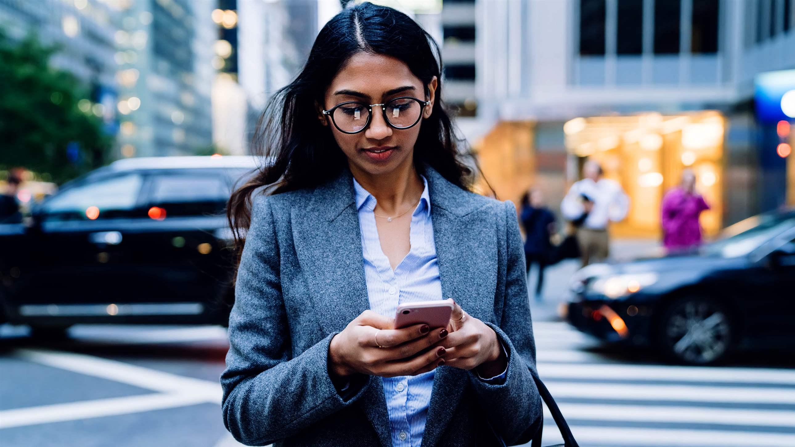 Asian business woman in glasses and gray jacket browsing in mobile phone while crossing road in New York City